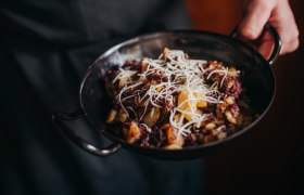 Eine Person h&auml;lt eine Pfanne mit Blunzengr&ouml;stl, garniert mit geriebenem K&auml;se.