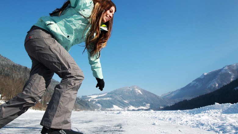 Eine Person eisläuft auf einem zugefrorenen See mit schneebedeckten Bergen im Hintergrund.