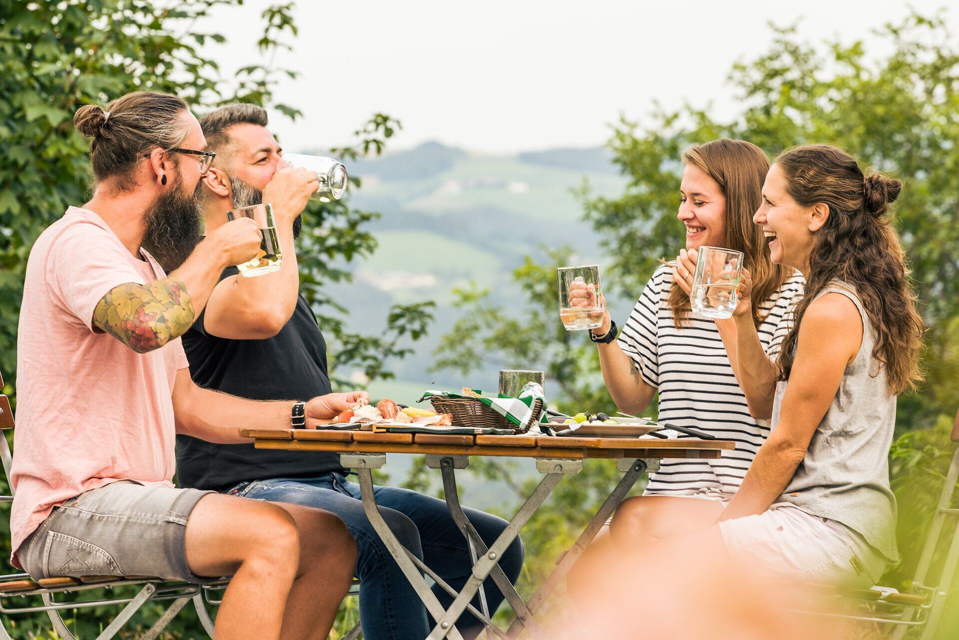 Zwei Frauen und zwei Männer sitzen sommerlich gekleidet und lachend an einem gedeckten Tisch beim Mostheurigen und halten große, gefüllte Gläser in ihren Händen. 
