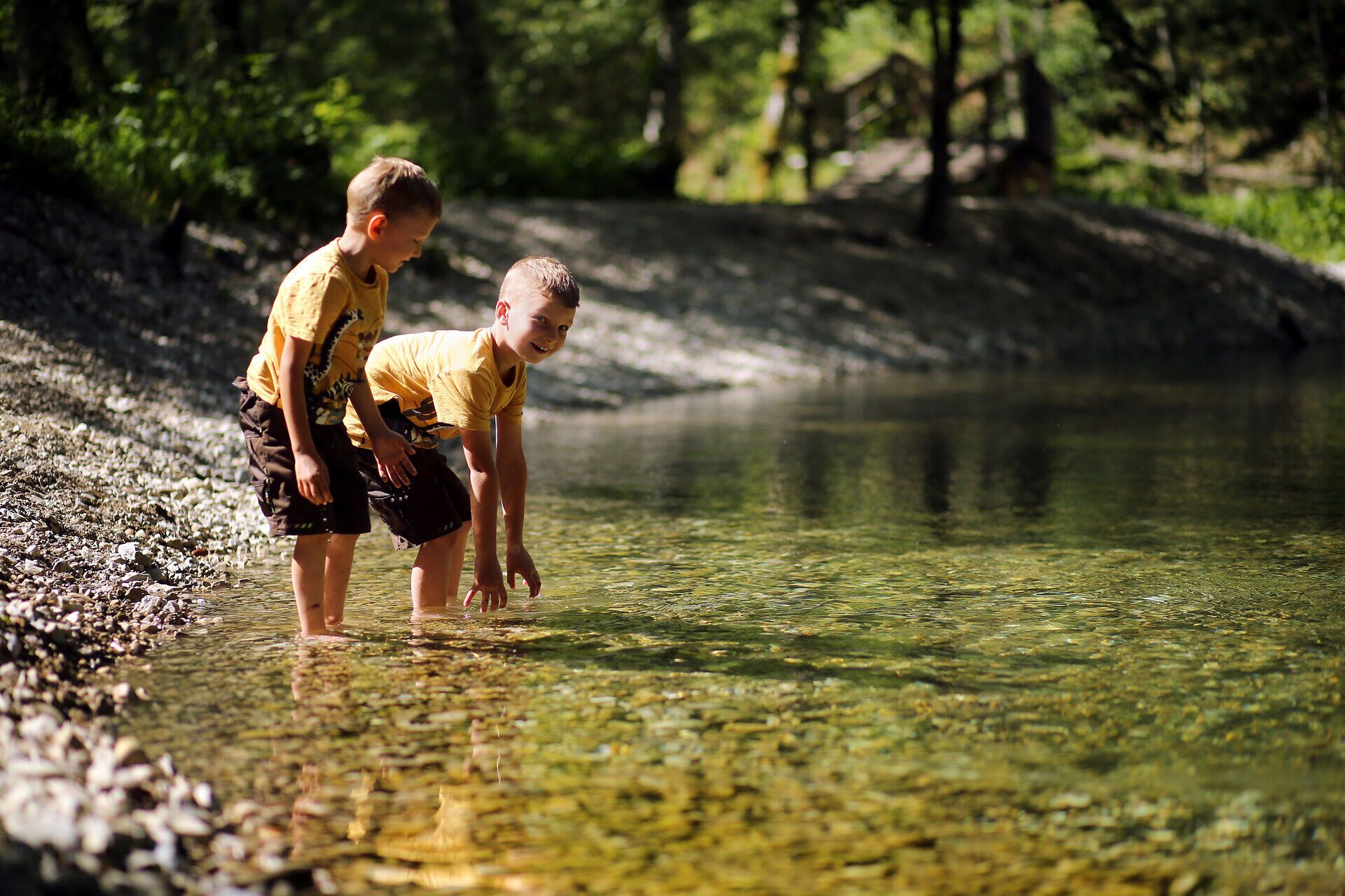 Zwei Kinder stehen am Ufer eines glasklaren Baches und tauchen neugierig ihre Hände ins Wasser. Umgeben von üppigem Grün und der sanften Stille der Natur, genießen sie den Sommer in den Bergen in vollen Zügen.