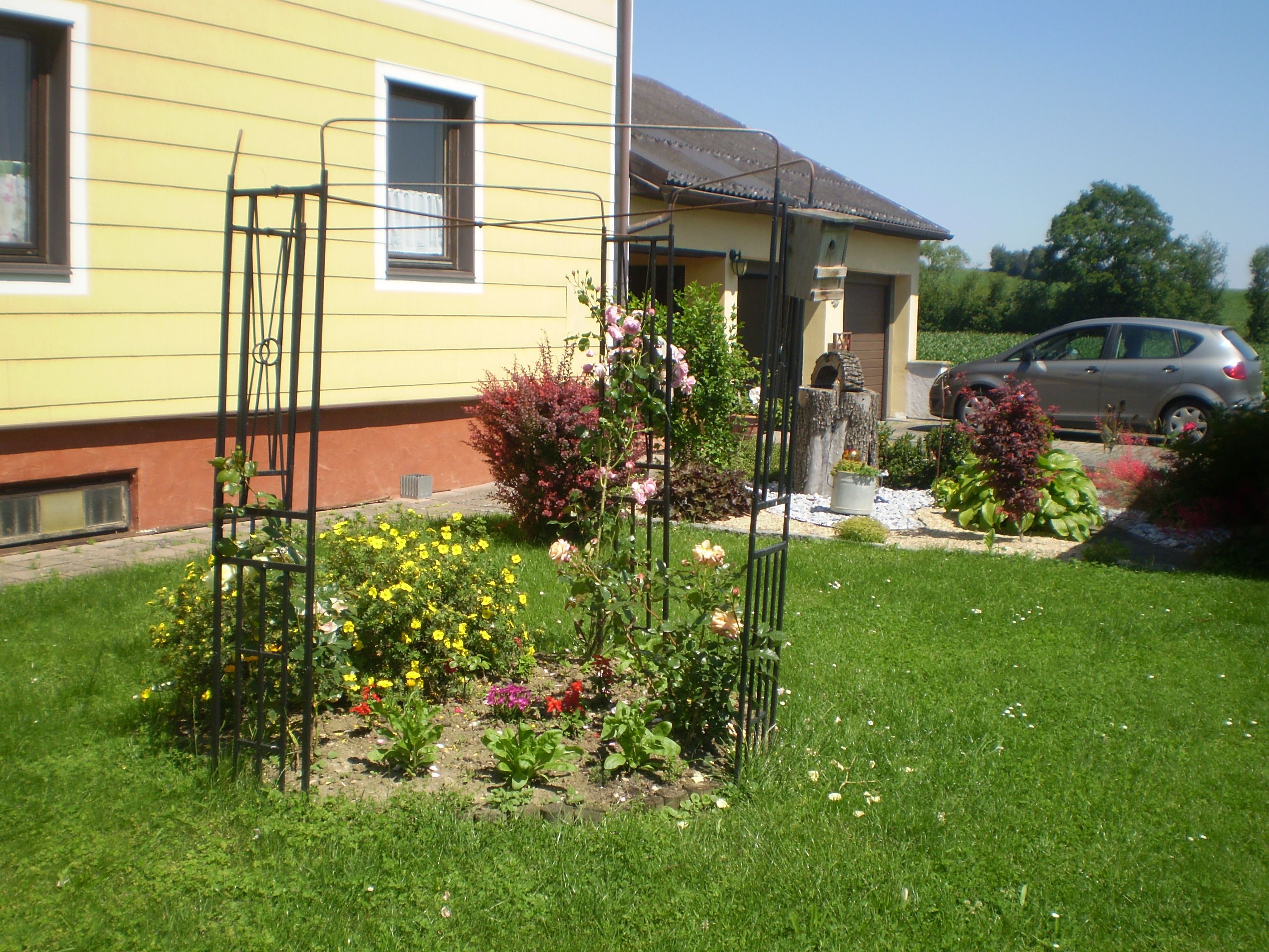 Ein gepflegter Garten mit Blumenbeet und Rankgerüst vor einem gelben Haus.