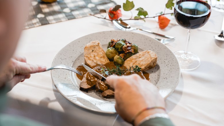 Person schneidet Fleisch auf einem Teller mit Knödeln und Rosenkohl, daneben ein Glas Rotwein.
