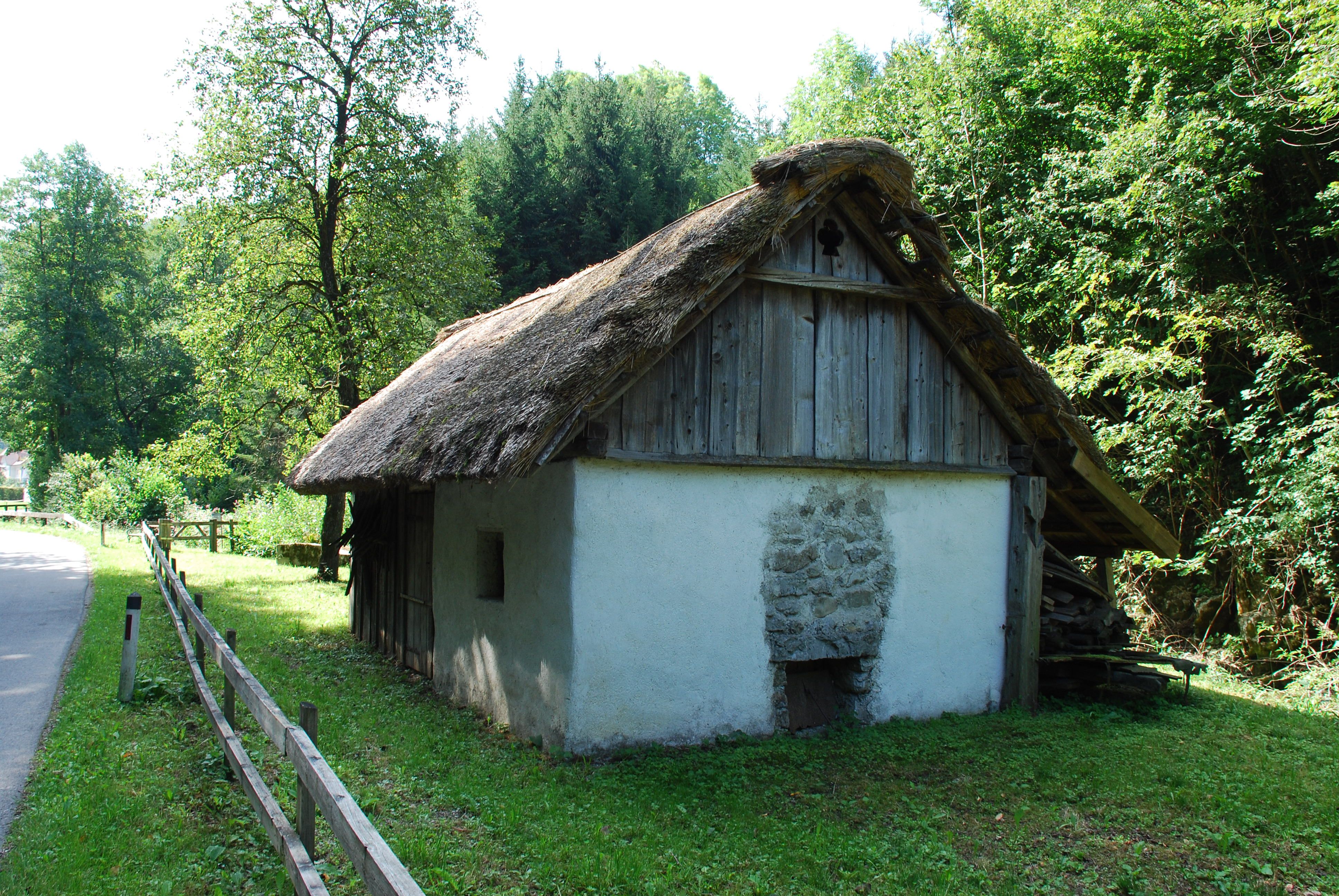 Ein traditionelles Dörrhaus mit Strohdach in einer ländlichen Umgebung, umgeben von Bäumen und Wiese.