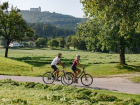 Ein Paar radelt entspannt durch die malerische Landschaft, umgeben von saftigem Gr&uuml;n und sanften H&uuml;geln. Die frische Luft und die strahlende Sonne schaffen eine einladende Atmosph&auml;re f&uuml;r einen aktiven Tag in der Natur. Im Hintergrund erhebt sich majest&auml;tisch ein Schloss, das die Sch&ouml;nheit der Umgebung perfekt erg&auml;nzt.