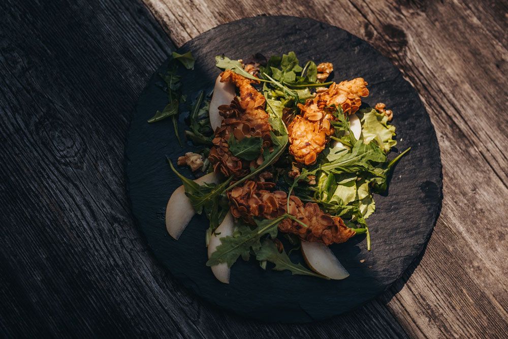 Hühnerfilet mit Mandelkruste auf Salat mit Birnenscheiben auf einem schwarzen Teller.
