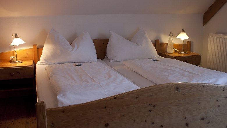 Gem&uuml;tliches Doppelbett in einem Landgasthofzimmer mit Holzrahmen und Nachttischen, beleuchtet von zwei Tischlampen.