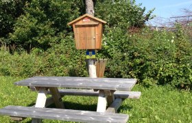 Picnic table next to a wayside shrine in a meadow with trees in the background.