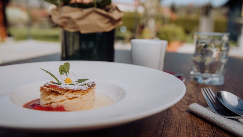 Ein Teller mit Milchrahmstrudel, garniert mit einem Gänseblümchen, auf einem Holztisch im Freien.