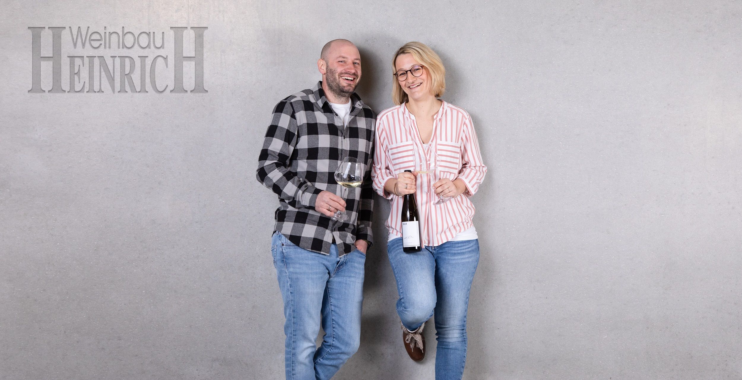 Zwei Personen stehen lächelnd vor einer grauen Wand mit Weingläsern in der Hand. Links steht ein Mann, rechts eine Frau. Oben links steht 'Weinbau Heinrich'.