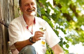 Mann sitzt draußen mit einem Glas Wein in der Hand, umgeben von grünen Pflanzen.