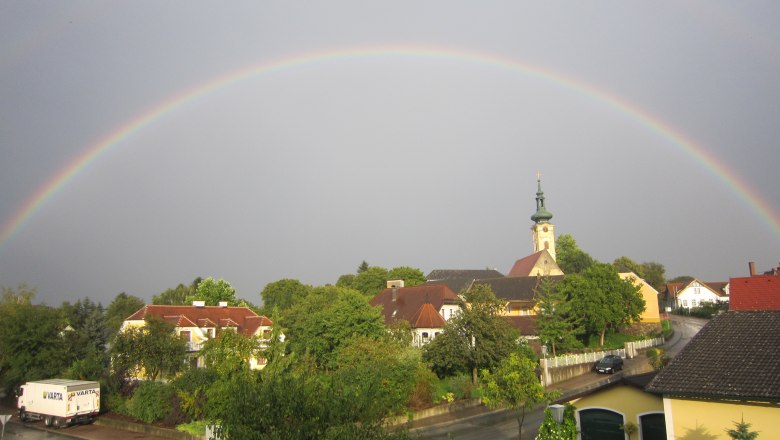 Ein Regenbogen spannt sich über ein Dorf mit Kirche und Häusern in Gerersdorf.