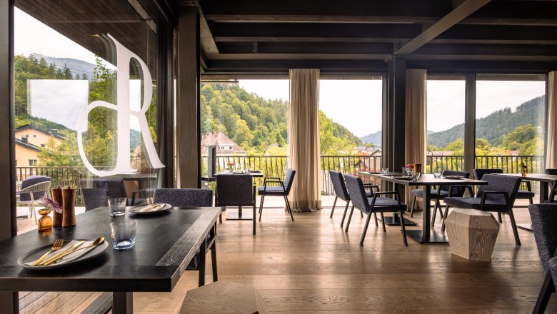 Innenansicht eines modernen Restaurants mit großen Fenstern und Blick auf bewaldete Hügel.