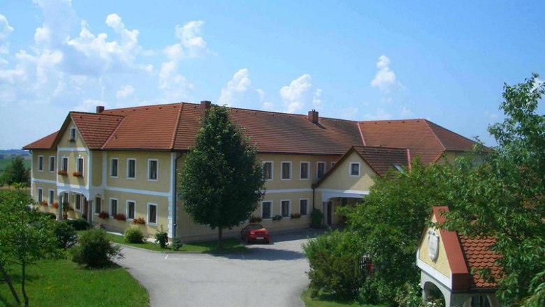 Großer Bauernhof mit gelben Wänden und roten Dächern, umgeben von Bäumen und einer Straße.