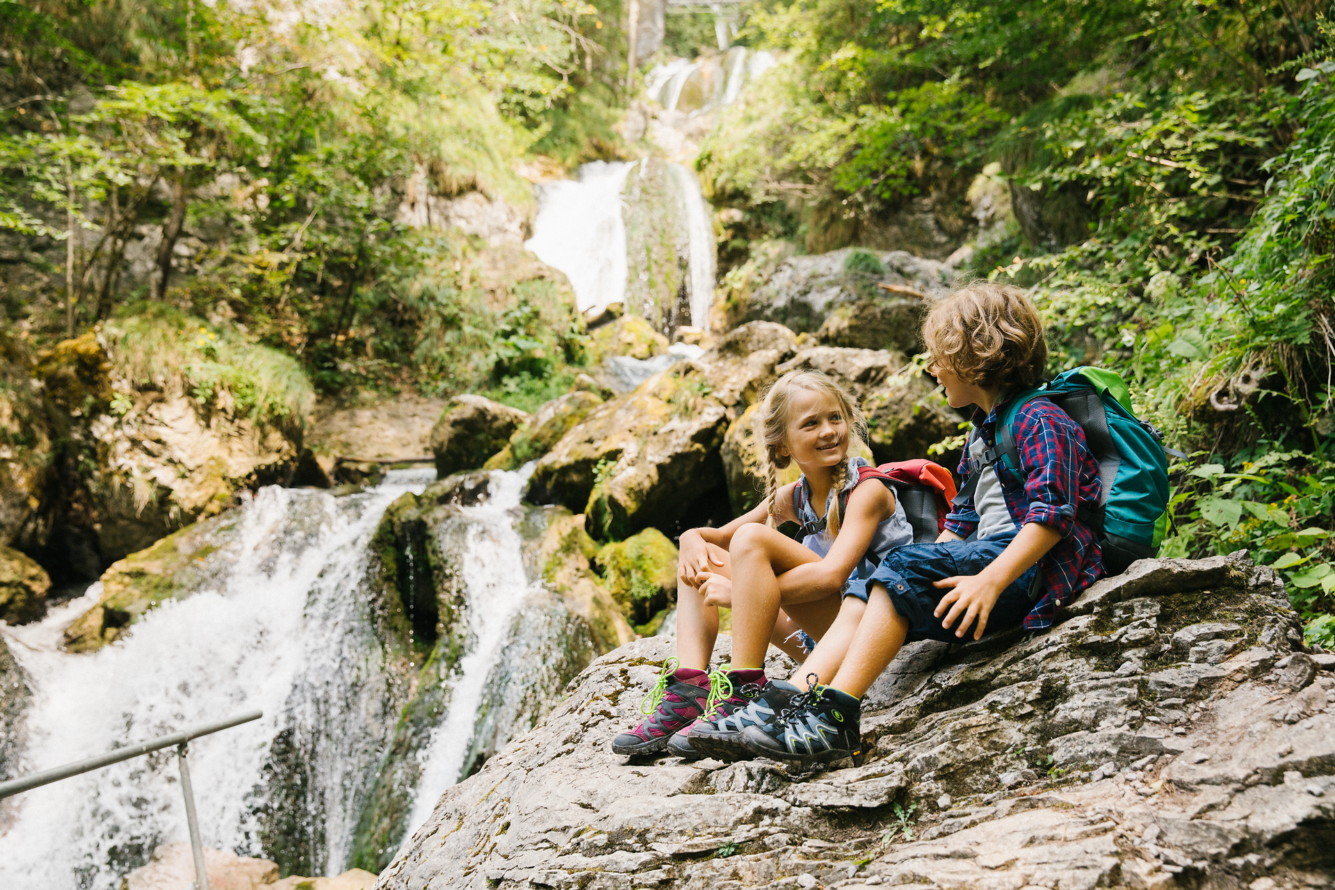 Zwei Kinder sitzen entspannt auf einem großen Stein und genießen die erfrischende Atmosphäre des Naturparks. Umgeben von üppigem Grün und dem sanften Rauschen des Wassers, strahlt die Szene pure Lebensfreude und Abenteuerlust aus.