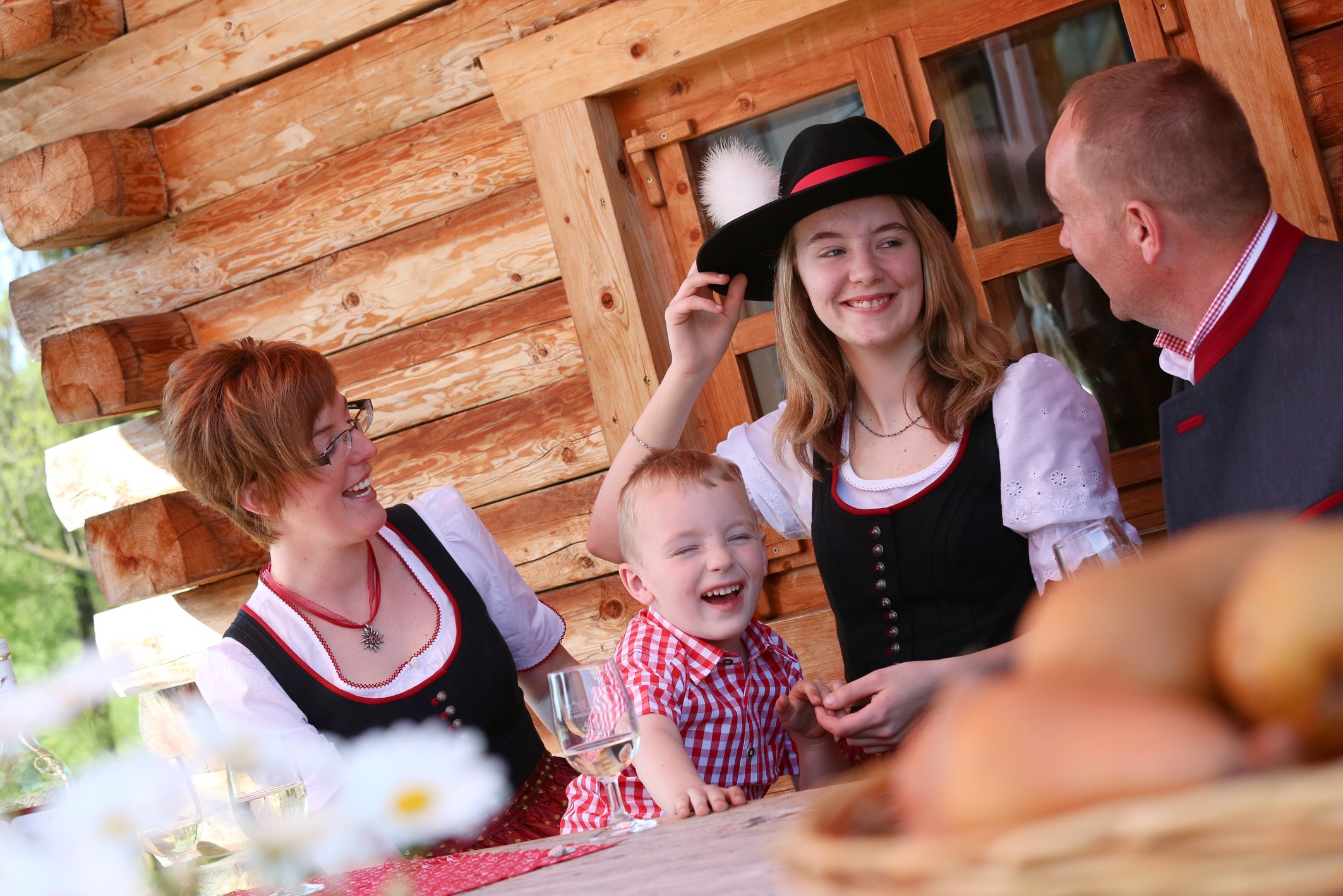 Familie in Tracht vor einer Blockhütte, lachend am Tisch.