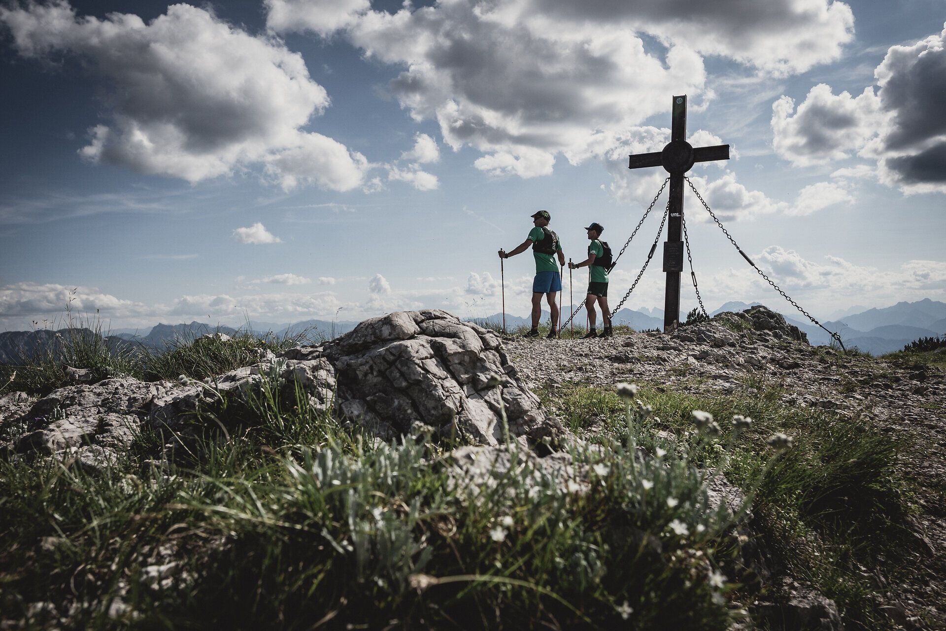 Zwei Wanderer genießen die frische Bergluft und die atemberaubende Aussicht, während sie sich dem Gipfelkreuz nähern. Umgeben von majestätischen Felsen und üppigem Grün, vermittelt die Szenerie ein Gefühl von Freiheit und Abenteuer. Der Sommer in den Bergen ist ein wahres Paradies für Naturliebhaber.