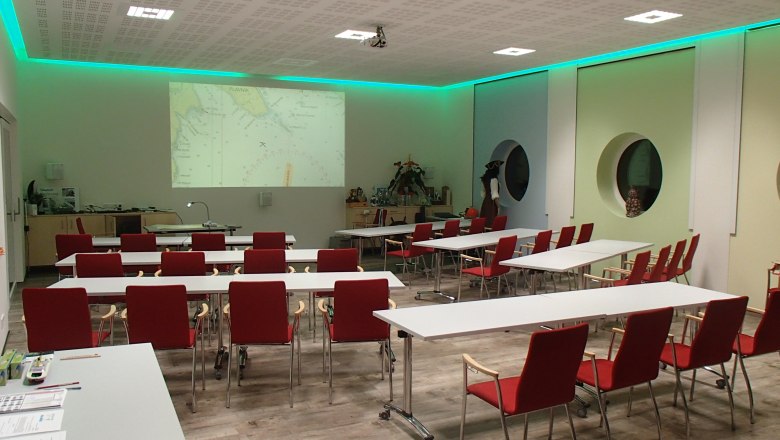 Training room with red chairs and white tables, projector shows nautical chart on the wall.