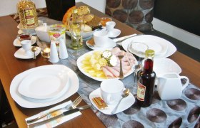 Frühstückstisch mit Geschirr, Aufschnitt, Brot, Müsli und Getränken.