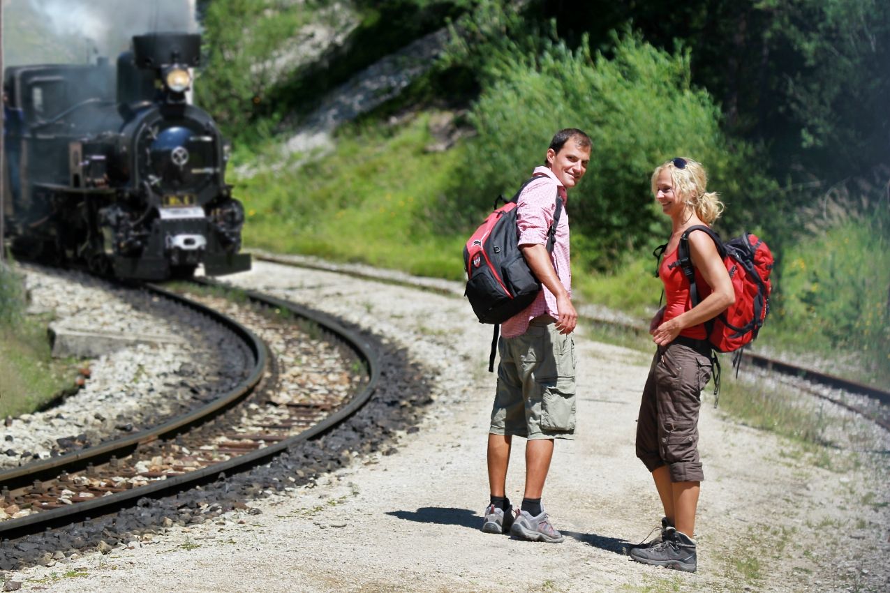 Zwei Wanderer stehen auf einem Schotterweg neben Bahngleisen, während eine Dampflok vorbeifährt.
