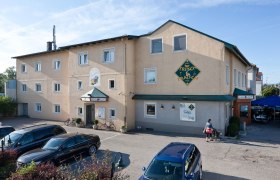 Außenansicht eines Gebäudes mit der Aufschrift 'Pension Schlafgut' und 'Café Trisch & Tratsch'. Autos parken davor, eine Person mit Kinderwagen geht vorbei.