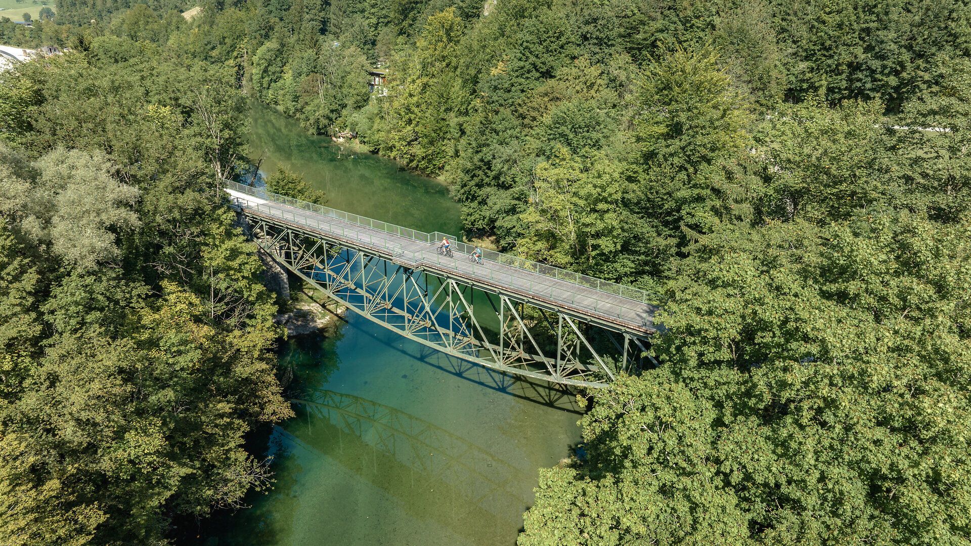 Eine malerische Brücke schwingt sich über das glitzernde Wasser, umgeben von üppigem Grün und majestätischen Bergen. Radfahrer genießen die frische Luft und die atemberaubende Aussicht auf die Ybbstaler Alpen, während sie die Ruhe der Natur erleben.