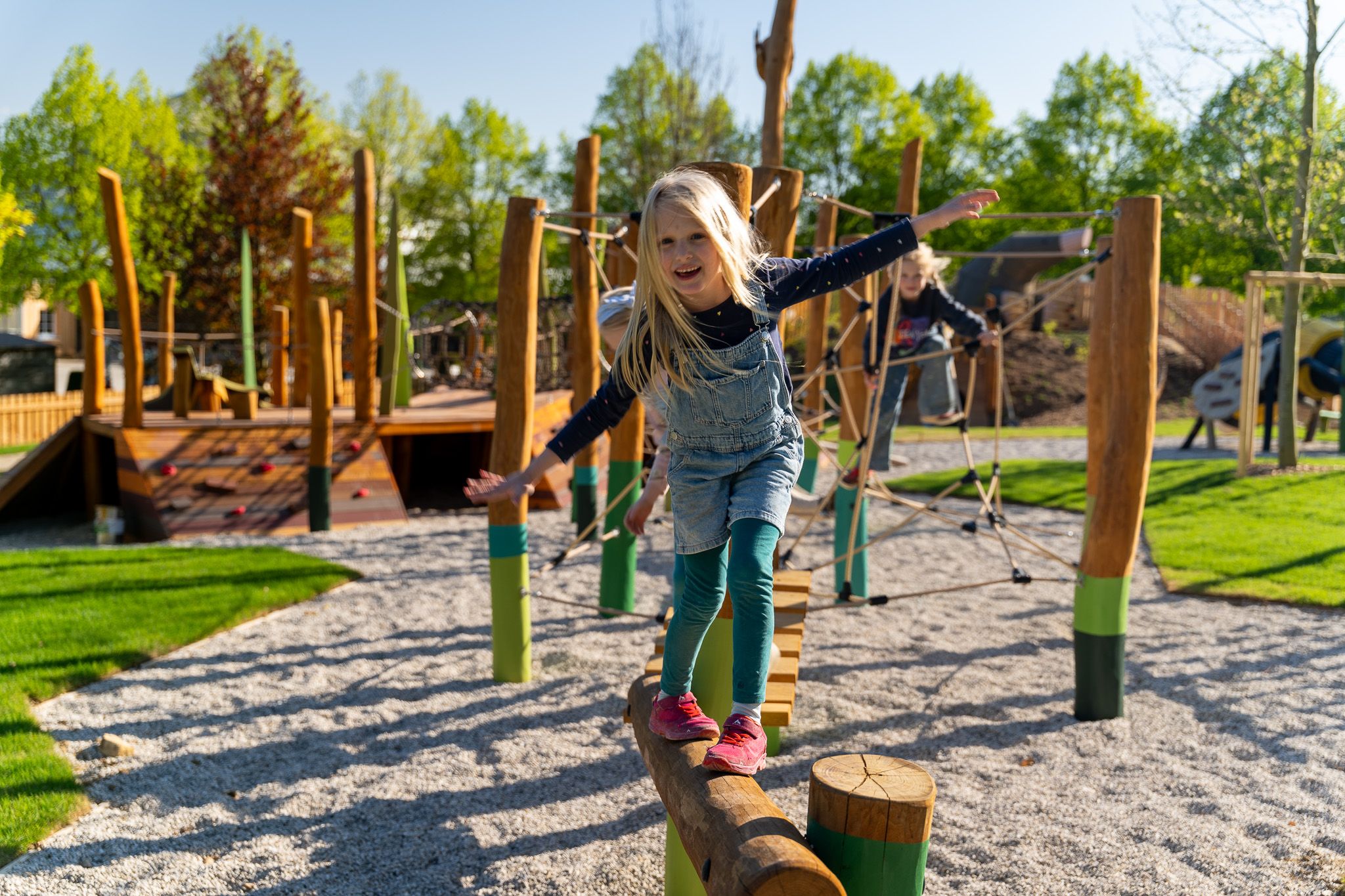 Ein Kind balanciert auf einem Holzstamm auf einem Spielplatz mit Klettergerüsten im Hintergrund.