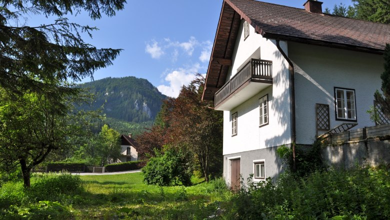Ein weißes Ferienhaus mit Balkon vor einer Berglandschaft und grünem Garten.