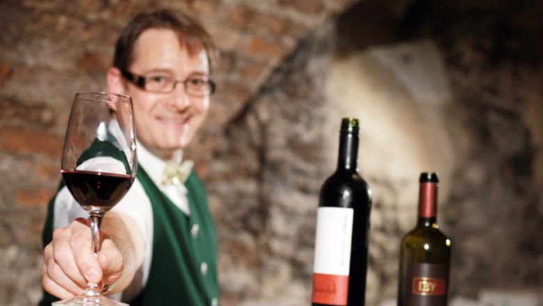 Ein Mann in traditioneller Kleidung bietet ein Glas Rotwein an, im Hintergrund zwei Weinflaschen.