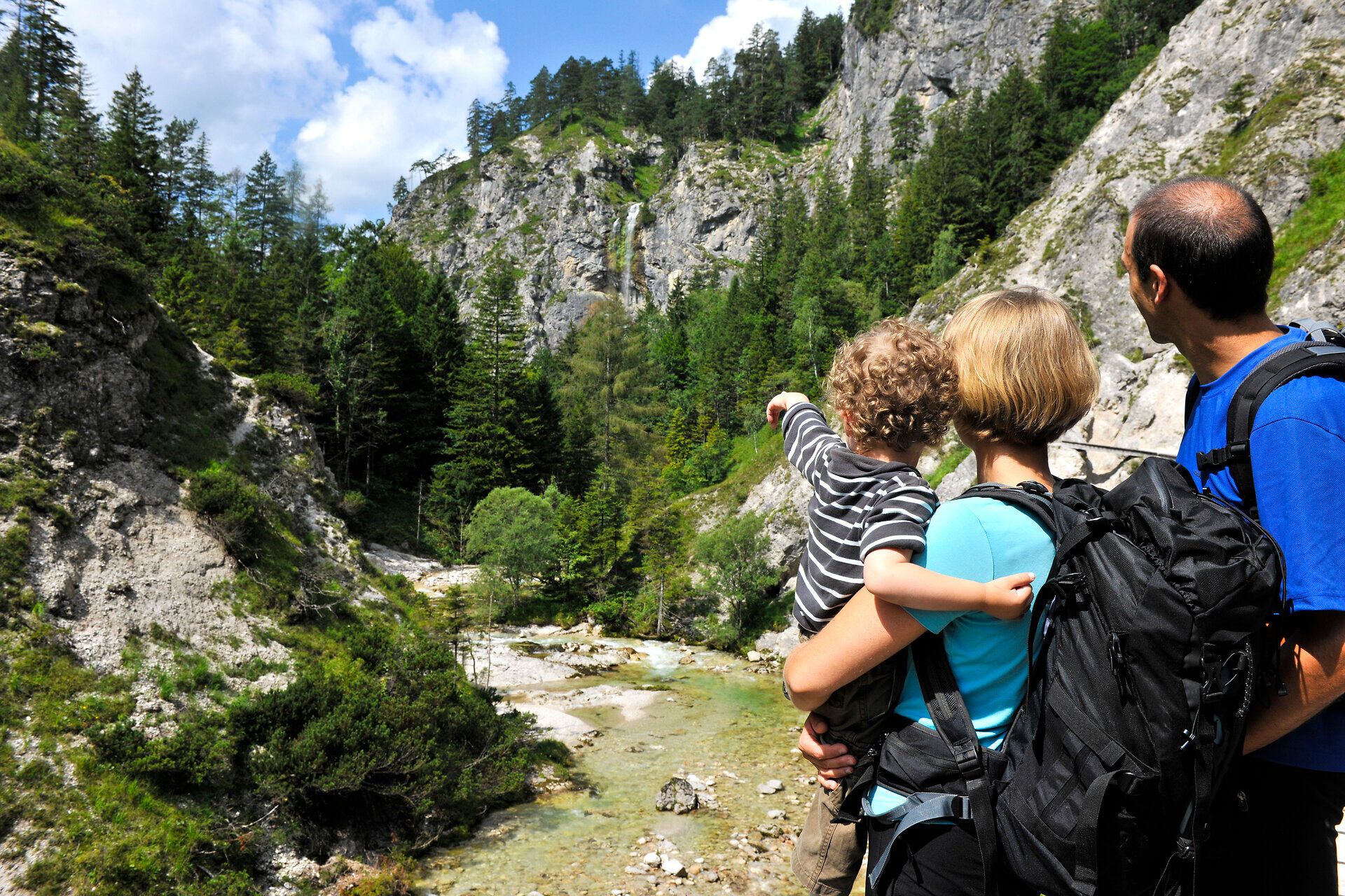Inmitten der majestätischen Berge genießen eine Familie die atemberaubende Aussicht auf die grüne Landschaft und den plätschernden Wasserfall. Die frische Bergluft und das sanfte Licht des Sommertages schaffen eine einladende Atmosphäre für unvergessliche Erlebnisse in der Natur.