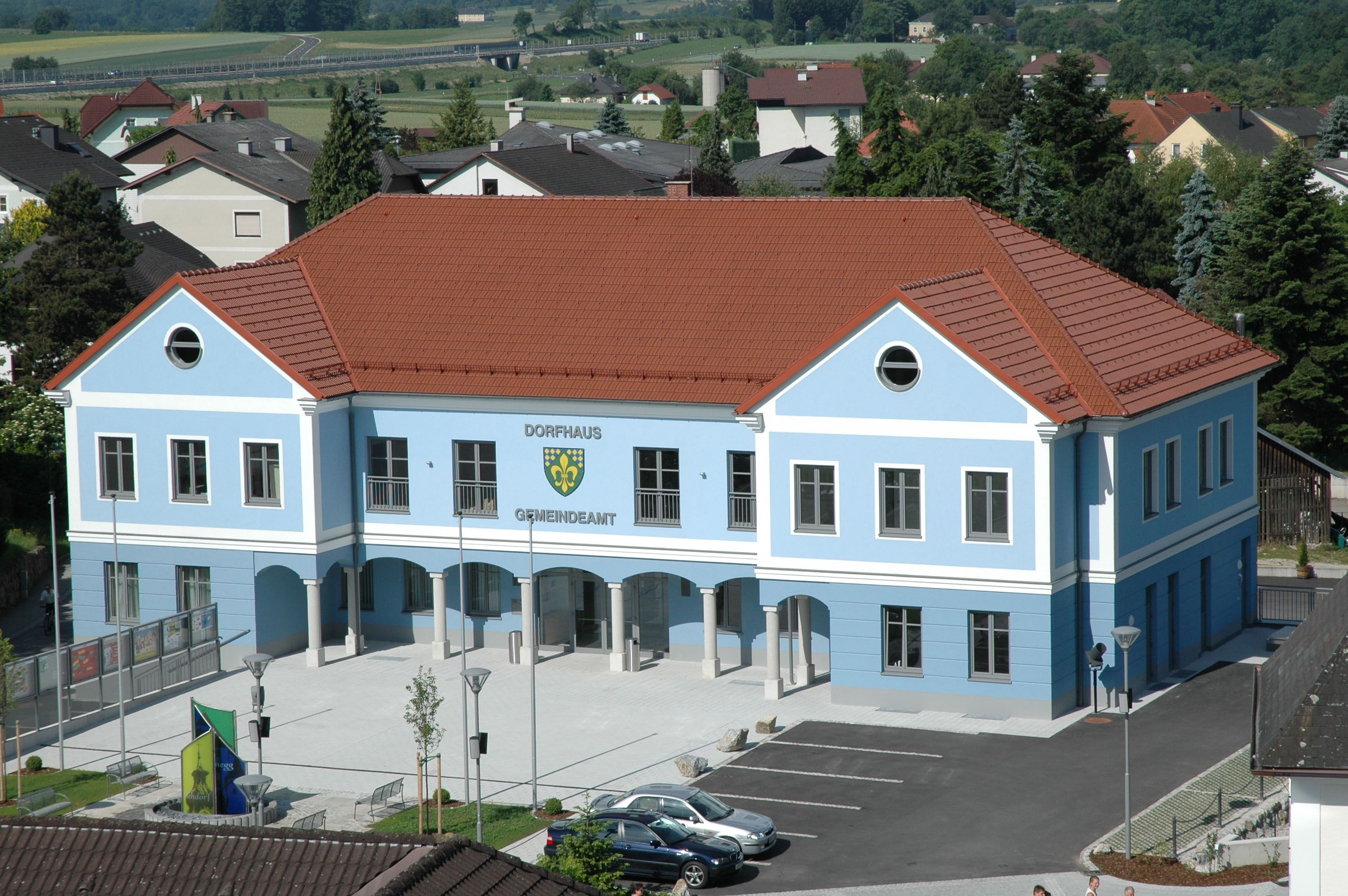 Blaues Gebäude mit rotem Dach, beschriftet als Dorfhaus Gemeindeamt, umgeben von Häusern und Bäumen.