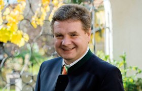 Ein l&auml;chelnder Mann in traditioneller Kleidung steht im Freien vor einem schmiedeeisernen Zaun mit gelben Bl&auml;ttern im Hintergrund.