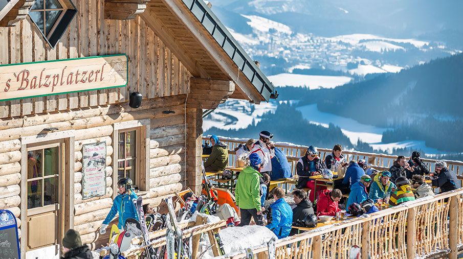 Menschen auf einer sonnigen Terrasse vor einer Berghütte mit Blick auf verschneite Berge.