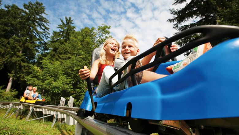 Zwei Kinder fahren lachend in einem blauen Sommerrodelbahn-Wagen durch einen Wald.