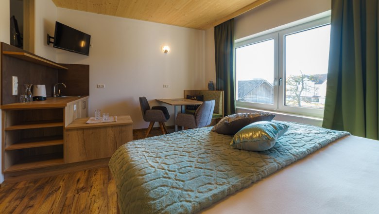 Gemütliches Hotelzimmer mit Holzboden, Doppelbett, kleinem Tisch und Stühlen, Fernseher und großem Fenster mit Ausblick.