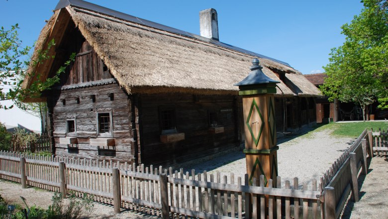 Traditionelles Holzhaus mit Strohdach und Holzzaun in ländlicher Umgebung.