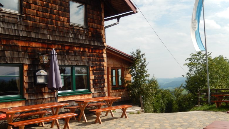 Holzh&uuml;tte mit Terrasse und B&auml;nken, Flagge weht im Wind.