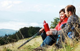 Ein Paar sitzt auf einer Wiese mit Wanderstöcken und einer roten Trinkflasche, im Hintergrund Berge und Wolken.