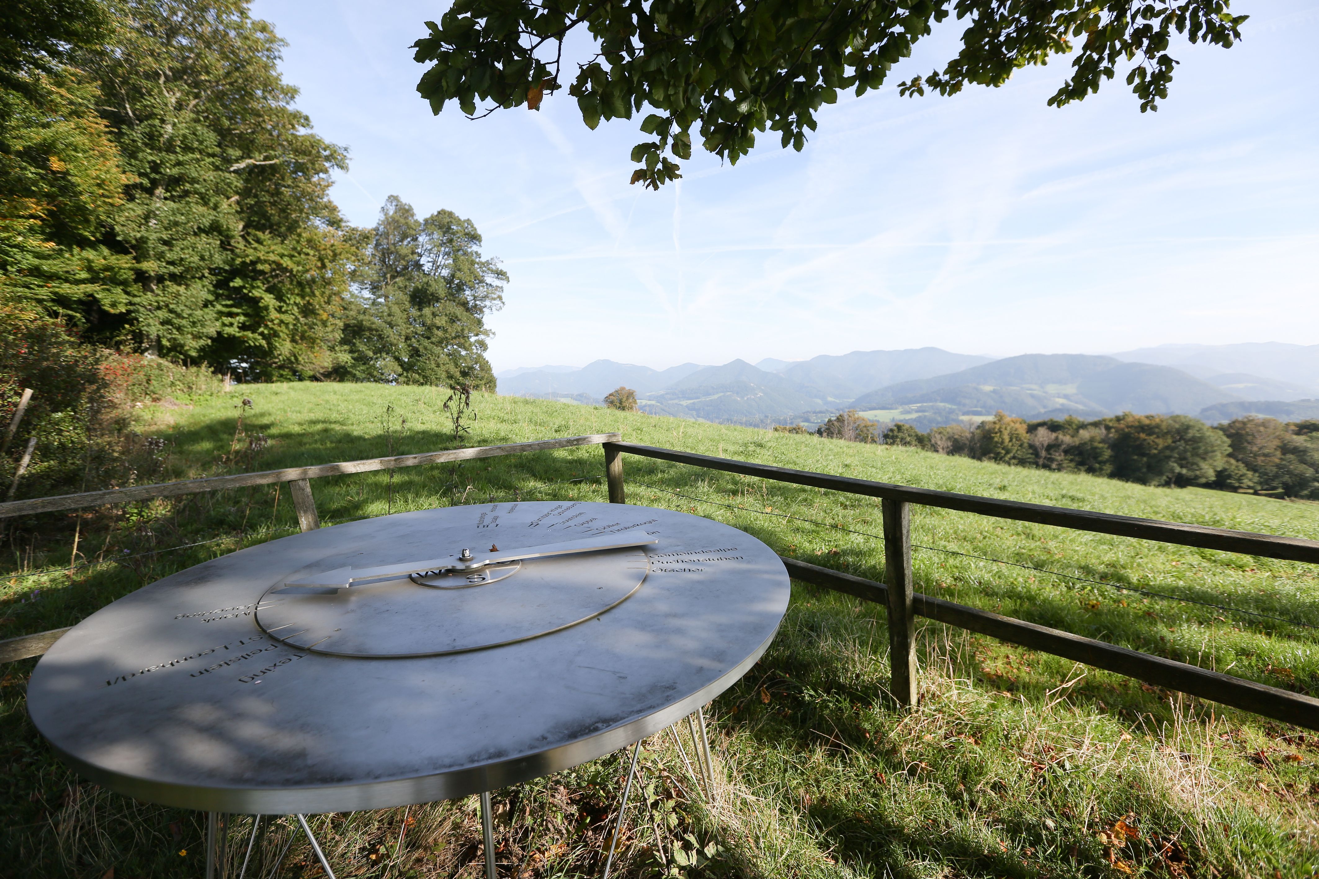 Panoramablick vom Bichlberg mit einer Metalltafel im Vordergrund und Bergen im Hintergrund.