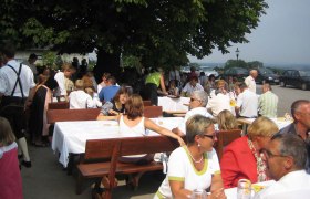 Menschen sitzen an langen Tischen im Freien unter einem Baum.