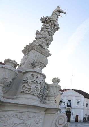 Nahaufnahme einer kunstvoll verzierten Säule mit Skulpturen und Ornamenten, im Hintergrund Gebäude und blauer Himmel.