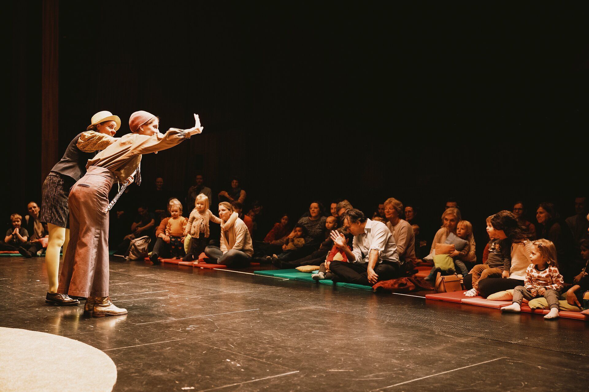 Kinder mit ihren Eltern im Publikum, im Vordergrund zwei Akteurinnen auf einer Bühne