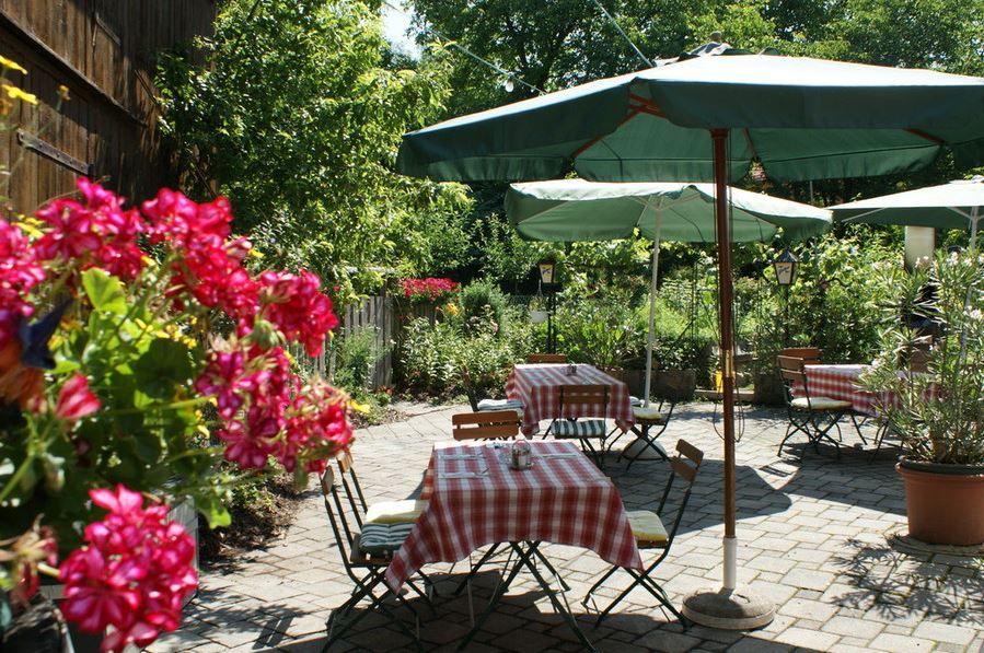 Ein gemütlicher Gastgarten mit rot-weiß karierten Tischdecken, grünen Sonnenschirmen und blühenden Blumen im Vordergrund.