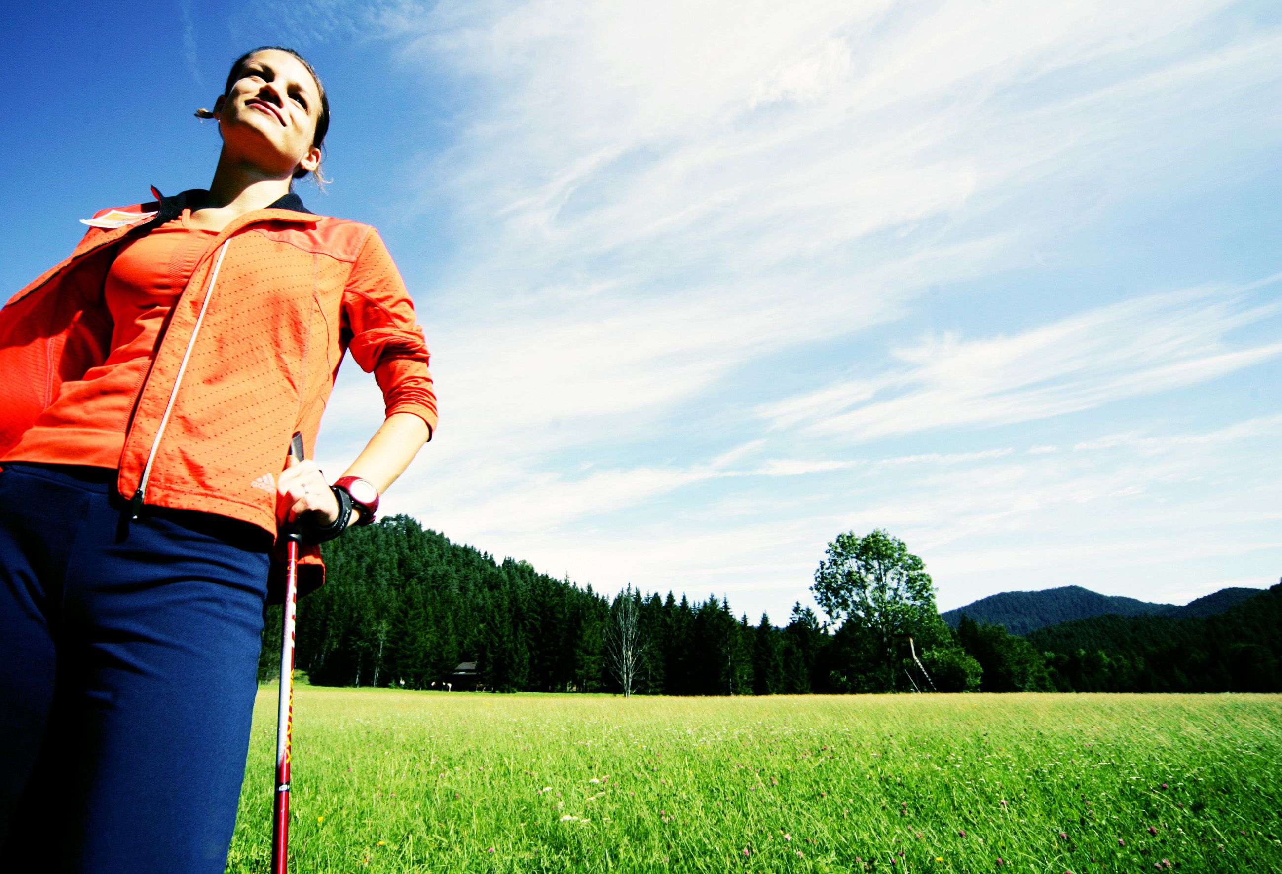 Frau in Sportkleidung mit Nordic Walking-Stöcken auf einer Wiese vor einem Wald.
