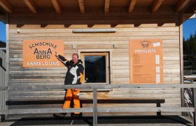 Ein Mann zeigt auf ein Schild der Schischule Annaberg vor einem Holzgeb&auml;ude.