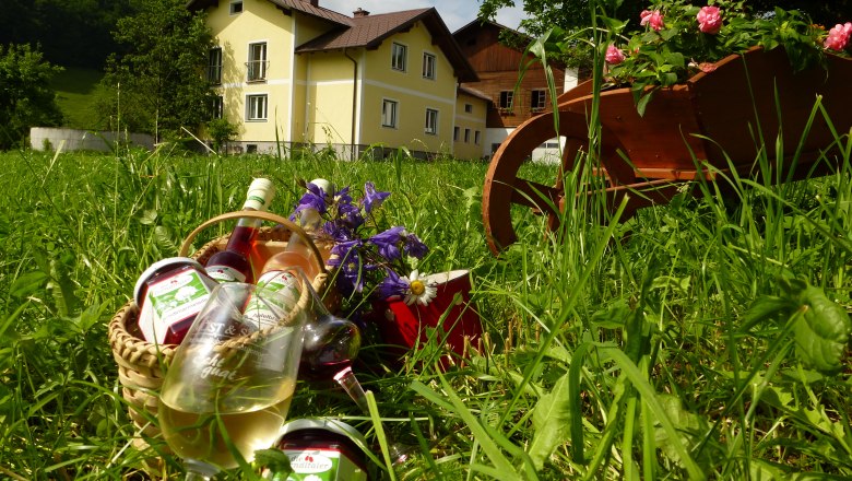 A yellow farmhouse in the background, in the foreground a basket with glasses and bottles on a meadow.