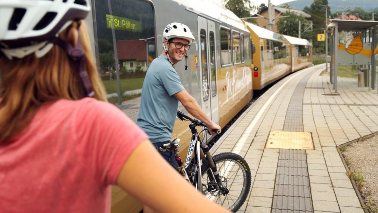 Zwei Personen mit Fahrradhelmen schieben ihre Räder am Bahnsteig, neben ihnen steht die Himmelstreppe auf den Gleisen.