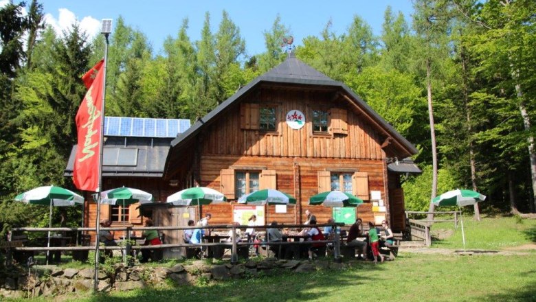 Holzh&uuml;tte im Wald mit Sonnenschirmen und Menschen auf der Terrasse.