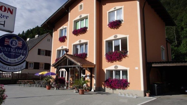 Ein dreistöckiges Gebäude mit orangefarbener Fassade und Blumenkästen vor den Fenstern. Ein Schild mit der Aufschrift 'Puntigamer' ist sichtbar.