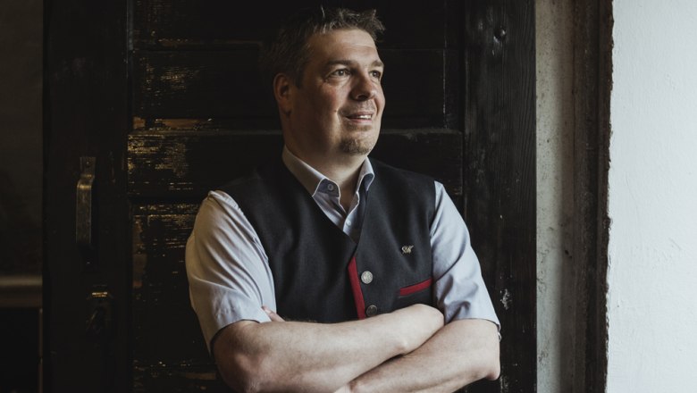 A man in traditional dress stands with his arms folded in front of a dark wooden door.