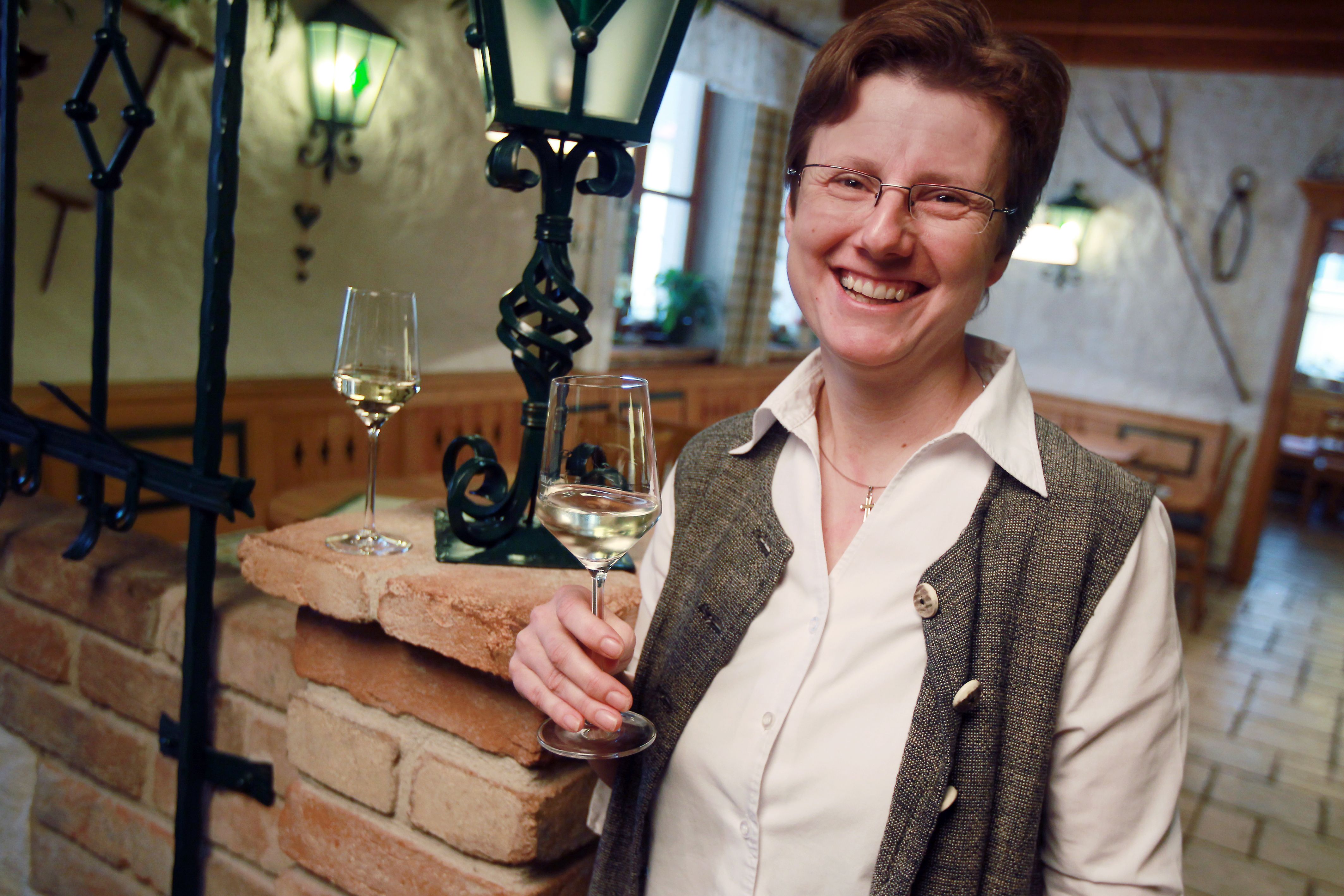 Eine lächelnde Person hält ein Glas Weißwein in einem gemütlichen Raum mit Backsteinmauer und Laterne.